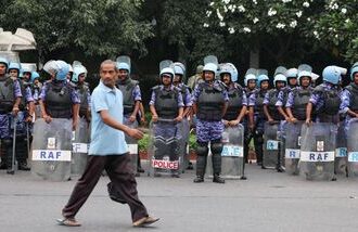 A man walks past members of the Rapid Action Force (RAF) standing guard ahead of a protest planned by the Cockroach Janta Party (CJP) seeking the resignation of India's Chief Election Commissioner Gyanesh Kumar, near the protest site in New Delhi
